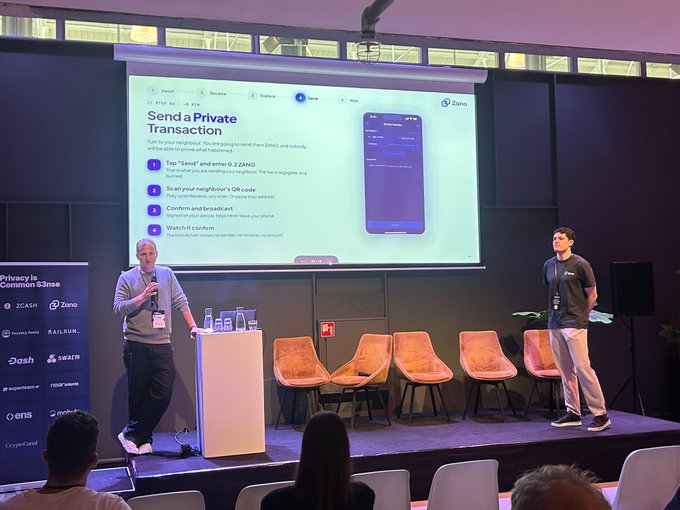 A photograph of a presentation stage where two men are standing in front of a large screen explaining how to 'Send a Private Transaction' using Zano, with logos for Zcash, Zano, Dash, and other crypto projects visible on the side.