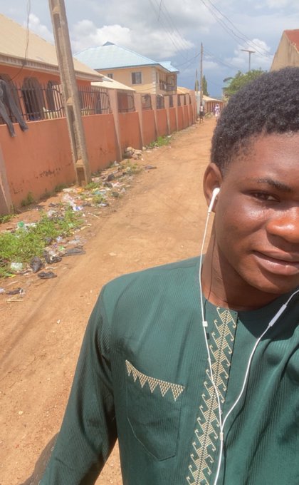 A young man wearing a green traditional embroidered tunic and white earphones takes a selfie on a dirt street, with orange-walled houses and a wall topped with barbed wire in the background.