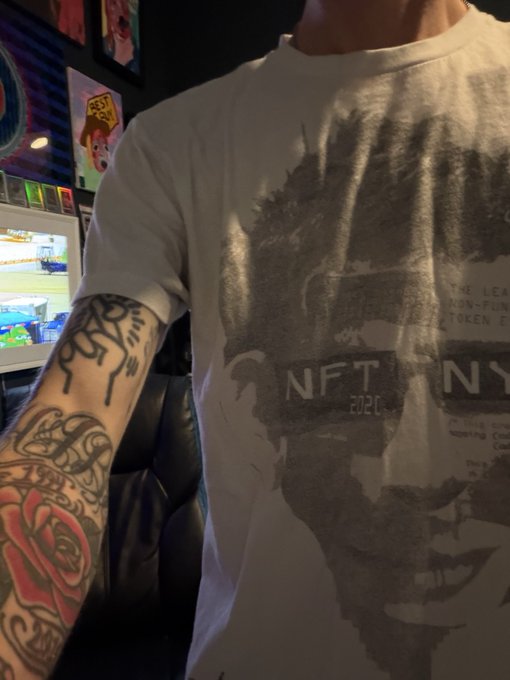 A person wearing a faded t-shirt featuring a Batman design with 'NFT NY 2020' text, displaying a heavily tattooed arm with red rose artwork.