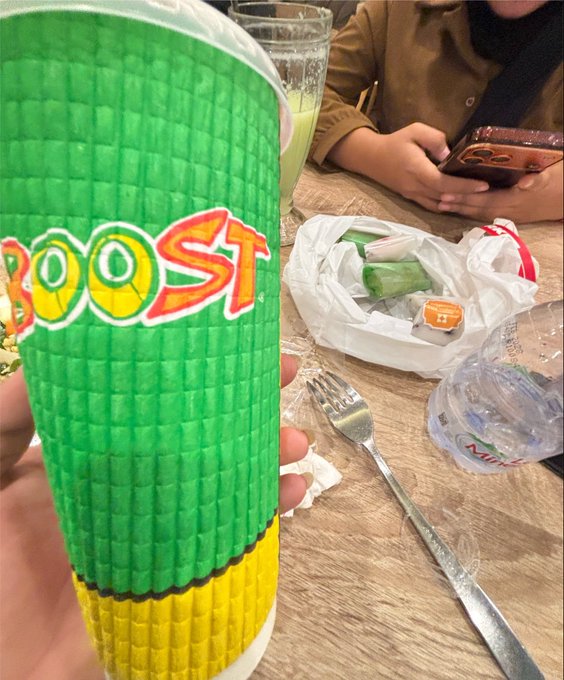 Close-up of a green piñata with the word 'BOOST' in colorful letters, held above a table with a fork, crumpled wrappers, and another person using a phone in the background.