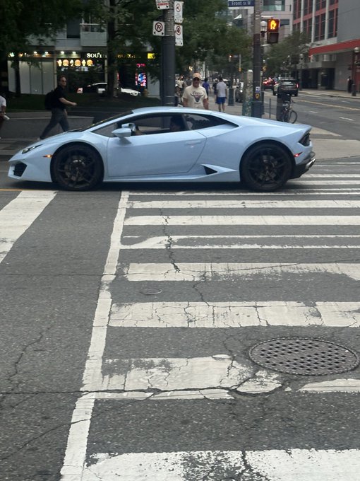 A light blue Lamborghini Huracán crosses a worn crosswalk on a city street, with pedestrians visible in the background near shops including a "Boka" storefront. The caption humorously suggests that the exotic supercar represents the "delusion" needed to survive a crypto bear market—dreaming of Lambos as one of the few coping mechanisms left alongside weed, Pokemon cards, Doginal Dogs, and coffee.