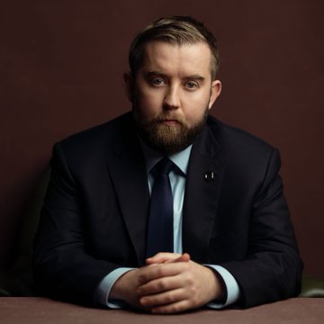 A portrait of a bearded man in a dark suit and blue tie, sitting with his hands clasped against a dark brown background.