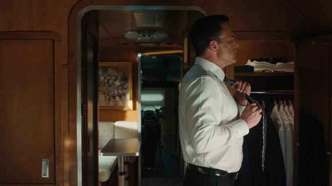A cinematic still of a man in a white dress shirt adjusting his tie in what appears to be a train compartment or wood-paneled room with hanging clothes.