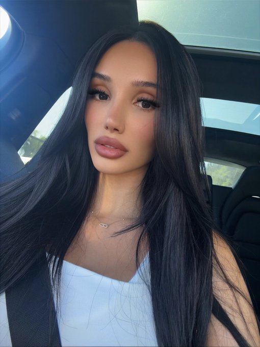 A selfie of a woman with long dark hair wearing a white top and a small necklace, sitting inside a car.