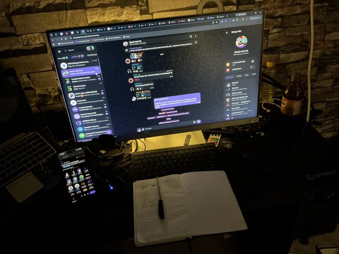 A dimly lit desk setup features a large monitor showing Discord and a phone with various app icons, accompanied by a mechanical keyboard, notebook, and pen.