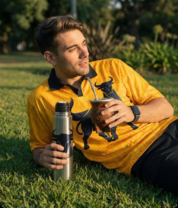 A man in a yellow sports jersey sitting on grass holding a traditional mate cup with a thermos, appearing relaxed outdoors.