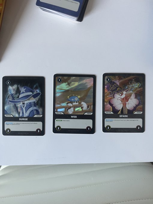 Three trading cards labeled 'SURGE', 'WEB', and 'SPARK' are laid out on a white surface next to a deck of cards, with WEB depicting a Smurf character on a pirate ship.