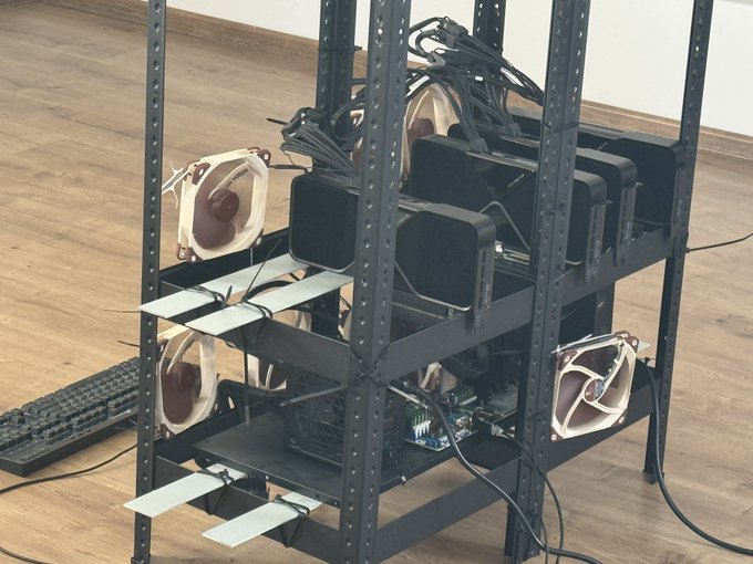 A DIY cryptocurrency mining rig sits on a metal shelving unit, featuring multiple GPUs with cooling fans, a motherboard, and tangled power cables — illustrating the homemade, self-built "GLM" (GPU Language Model) setup the author references in their post as they poke fun at the overhyped "RSI" (Robotic/Recursive Self-Improvement) buzzword.
