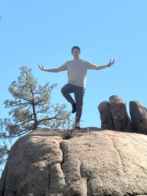A man stands balanced on one leg with arms outstretched in a yoga pose atop a large rock formation under a clear blue sky, with a pine tree visible on the left.