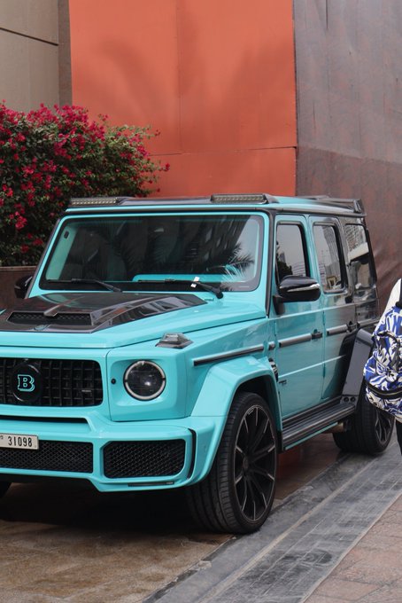 A bright turquoise Mercedes-Benz G-Wagon (Brabus edition) parked near a terracotta-colored wall with pink flowers.
