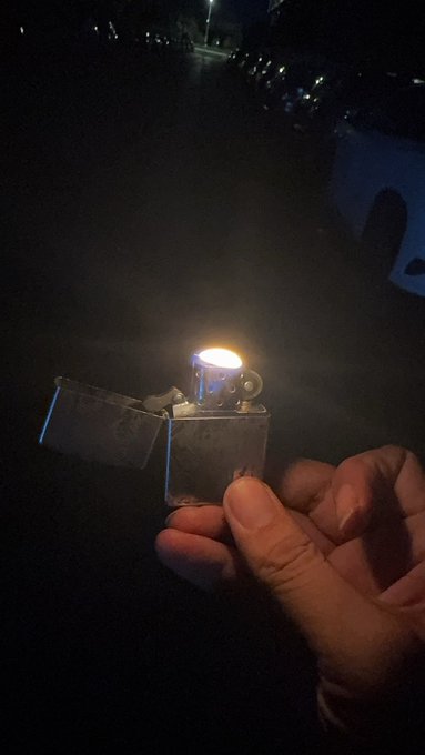 A hand holds a lit lighter in a dark outdoor setting, illuminating the flame against a night background.