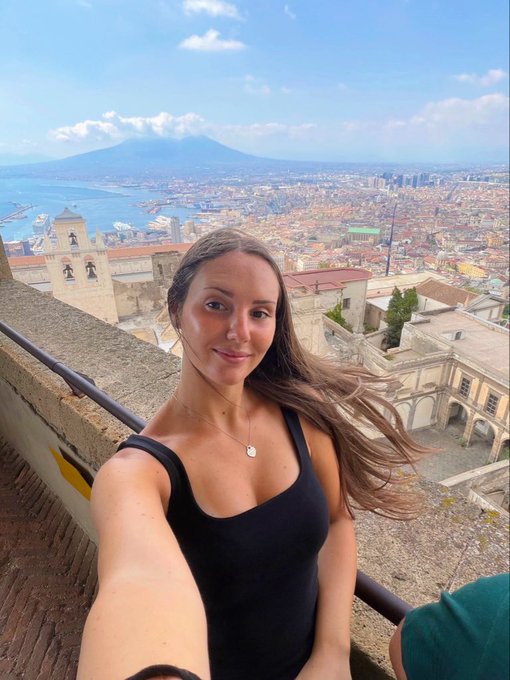 A selfie of a young woman with long brown hair wearing a black tank top, taken from a high vantage point overlooking a coastal city with a large volcano, likely Mount Vesuvius, in the background.
