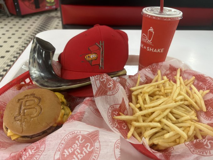 A meal at Steak 'n Shake featuring a cheeseburger with a Bitcoin logo stamped on the bun, fries, and a milkshake on a tray, alongside a red baseball cap.