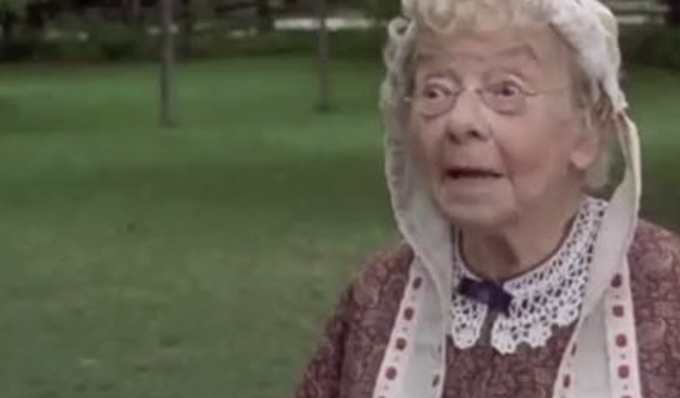 The image features an older woman wearing a white hat with lace trim and a red and white dress. She appears to be posing for a photo or video, possibly as part of a promotional campaign. The background shows a grassy field, which could suggest that she is in a park or outdoor setting. There are also trees visible in the scene, adding to the natural ambiance. The image includes text that reads "if shipping on Solana is cool, consider me Miles Davis", possibly indicating an association between the woman and the musician Miles Davis.