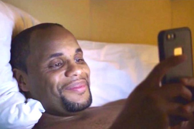 A man reclines on a white pillow in a warmly lit room, smiling while looking at and holding a smartphone.