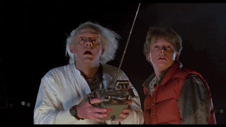 The image features two people, one wearing a white shirt and another wearing an orange vest. The person in the white shirt has their face covered with white hair and appears to be holding a small object or device. The other person, dressed in an orange vest, is looking up at the person in the white shirt. The background of the image is dark, which contrasts with the two individuals' appearances. There are no texts or additional objects present in the image.