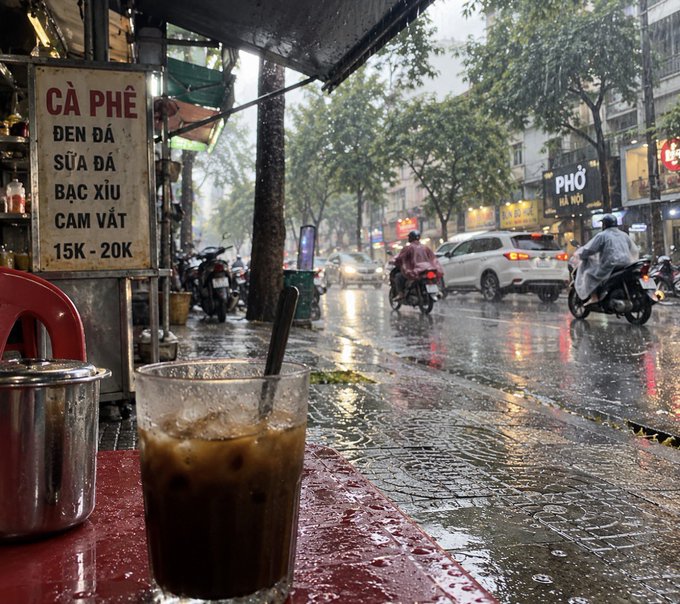 A glass of iced Vietnamese coffee with a black straw sits on a red table at a street-side café on a rainy day. A sign reads 'CÀ PHÊ ĐEN ĐÁ, SỮA ĐÁ, BẠC XỈU, CAM VẮT 15K - 20K' with motorbikes and cars passing by on the wet street.