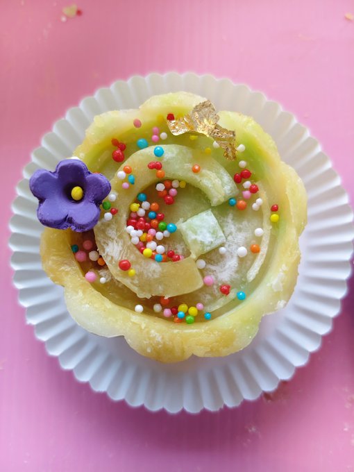 A close-up of a decorative rose-shaped dessert in a white paper cup on a pink background, topped with colorful sprinkles, gold leaf, and a small purple flower.