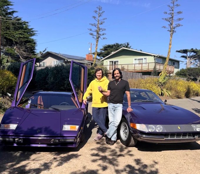 Two men pose between two purple sports cars—a Lamborghini with scissor doors open and a Ferrari—in front of a residential house.
