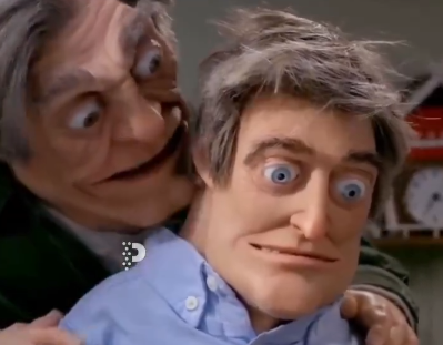 Two rubber hand puppets, one appearing as an older man grabbing from behind another man with wide blue eyes, are shown in a classic ventriloquist act.