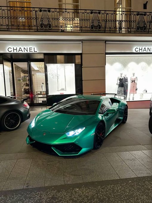 A green Lamborghini Huracán parked on a street at night in front of a luxury Chanel boutique with illuminated window displays.