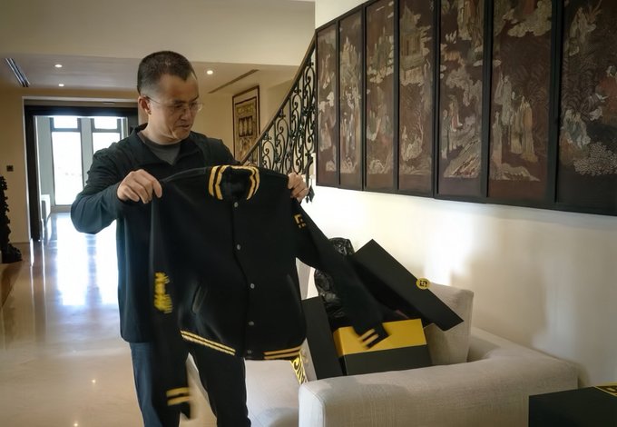 A man wearing glasses holds up a black and yellow varsity-style jacket, displaying it in an elegant home interior featuring decorative Asian artwork panels and ornate iron stair railings. The image aligns with the post's theme of CZ's understated personal style, showing him in casual attire that contrasts with the refined, culturally rich surroundings of his Dubai residence.