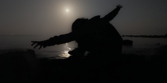 A silhouette of a person crouching with arms outstretched is captured against a dark, moonlit body of water.