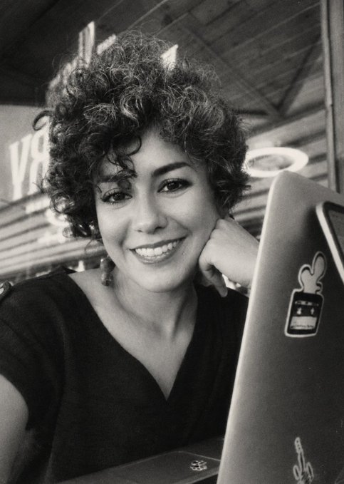 A black-and-white photograph of a smiling woman with curly hair posing next to a laptop adorned with stickers.