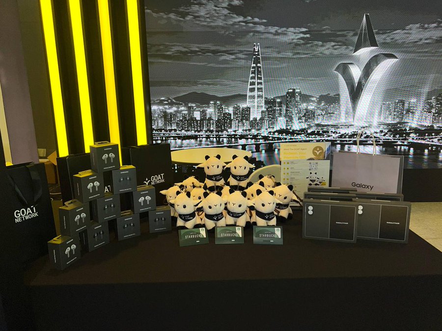 A promotional display featuring GOAT Network merchandise, Samsung Galaxy products, Starbucks gift cards, and plush goat toys, with a black and white Seoul cityscape backdrop showing Namsan Tower and City Hall.