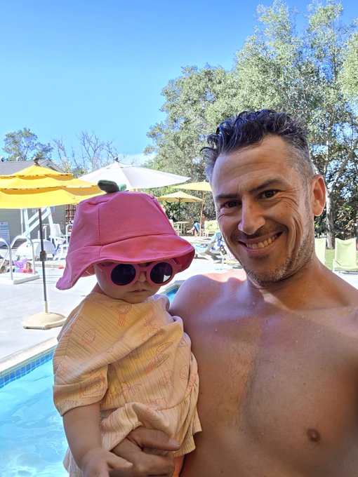 A shirtless smiling man holds a small child wearing a pink bucket hat, sunglasses, and a peach-colored outfit by a pool, with yellow umbrellas and trees in the background.