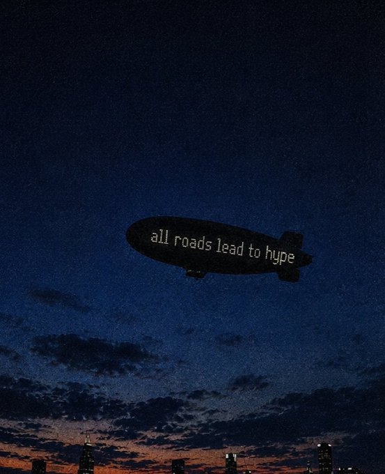 Nighttime photograph of a blimp flying over a city skyline with the text 'all roads lead to hype' displayed on its side.