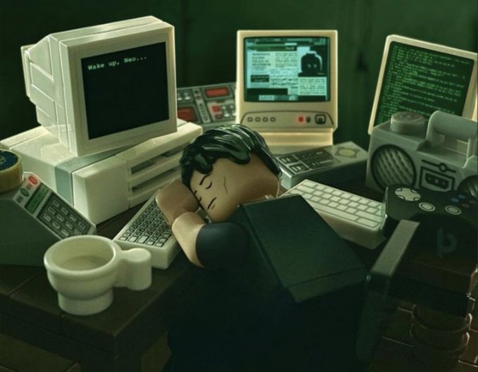 A miniature figure sleeps exhausted at his desk surrounded by vintage computers, one displaying "Wake up, Neo..."—a playful "gm" post referencing The Matrix, humorously wishing someone a good morning while suggesting they're still half-asleep in the simulation.