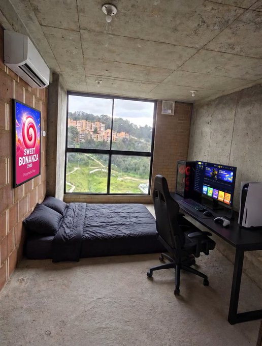 A bedroom with exposed concrete walls and ceiling, featuring a large window with a green landscape view, a bed, a gaming desk with dual monitors and PC, and a 'Sweet Bonanza' poster on the wall.