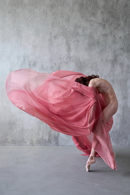 A ballet dancer in a flowing pink dress bends backward with the fabric dramatically draped over her head, posed on pointe against a textured gray wall.
