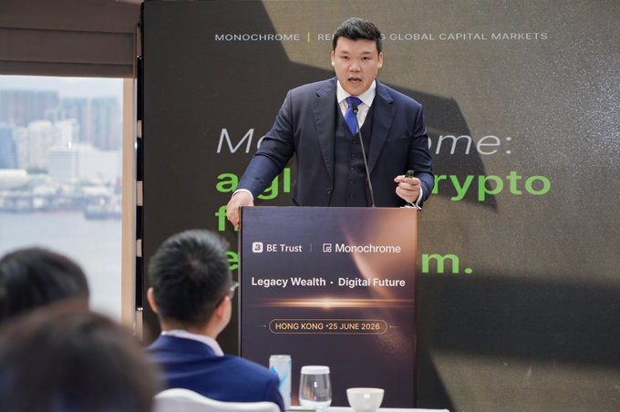 A man in a suit speaking at a podium during the 'Monochrome | Redefining Global Capital Markets' event in Hong Kong on June 25, 2026, with a banner reading 'Legacy Wealth • Digital Future' and 'BE TRUST'.