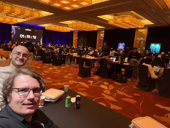 Two men taking a selfie at what appears to be a competitive gaming or hacking event, with a timer showing '01:51:12' displayed on a large screen in the background.