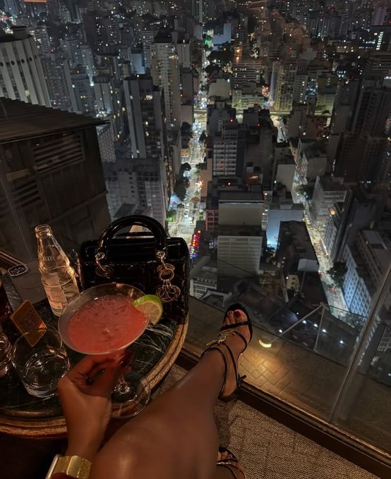 First-person perspective photo of a person holding a pink cocktail with lime while sitting at a high-rise bar overlooking a sprawling cityscape at night, with a luxury handbag visible.