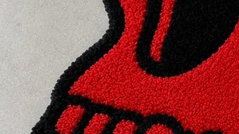 A close-up shows the textured, tufted surface of a red and black floor mat or rug with curved black design elements on a light gray background.