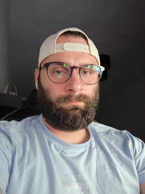 The image features a man with glasses wearing a white baseball cap sitting down. He is looking directly at the camera, with his left hand resting on his chin and right hand placed on his hip. The background seems to be a dark room or an indoor space with some light coming in from the top left corner.