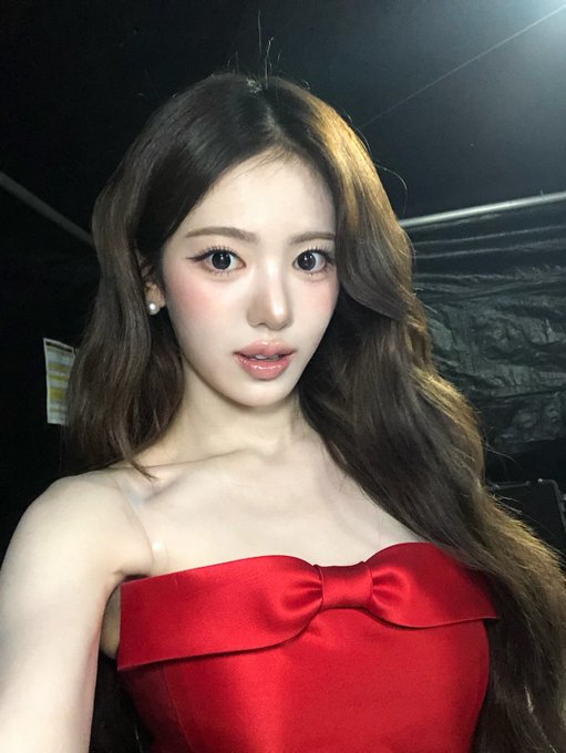 A young woman with long, wavy brown hair wearing a strapless red satin dress with a bow detail stands in a dimly lit setting.