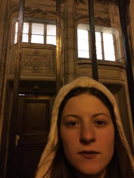 A woman wearing a hooded sweatshirt takes a selfie in front of what appears to be a synagogue, identifiable by the Star of David above the entrance. The image accompanies a post claiming the person in the photo was responsible for doxxing Bastille and suggests she deserves the same treatment in retaliation.