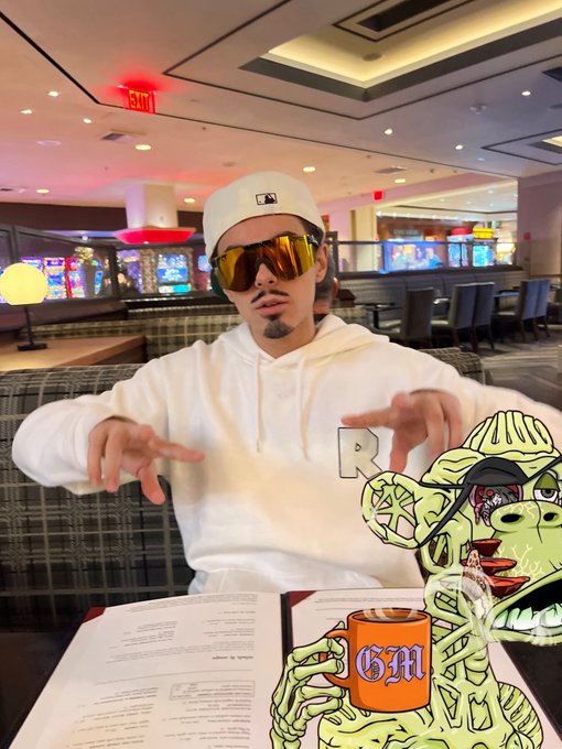 A young man wearing a white cap, yellow-tinted sunglasses, and a white hoodie sits in what appears to be a casino, with a cartoon Pepe frog holding a beer mug overlaid on the right side.