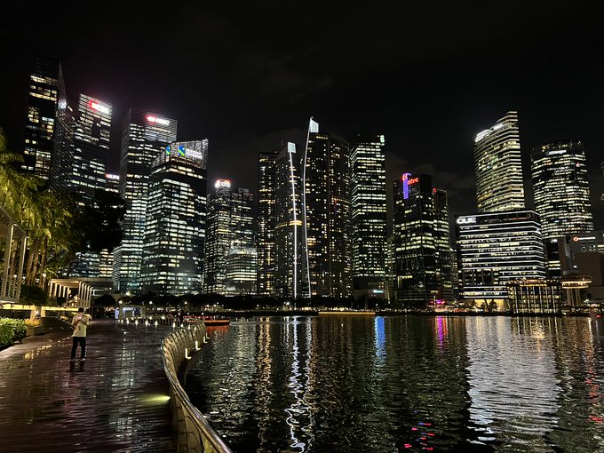 Nighttime cityscape showing illuminated skyscrapers reflected on the water of Marina Bay in Singapore, featuring palm trees along the waterfront promenade.