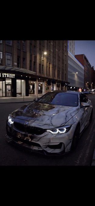 A white marble-patterned BMW coupe with illuminated angel-eye headlights is parked on a city street at night, with a storefront sign partially reading 'STREET' visible in the background.
