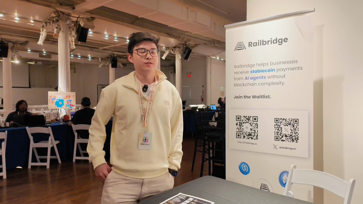 The image features a man standing next to a table with a sign that says "railbridge" on it, as well as a QR code. The man appears to be wearing glasses and has his hands in his pockets. There are other people present in the background, but they are not the main focus of the scene.
