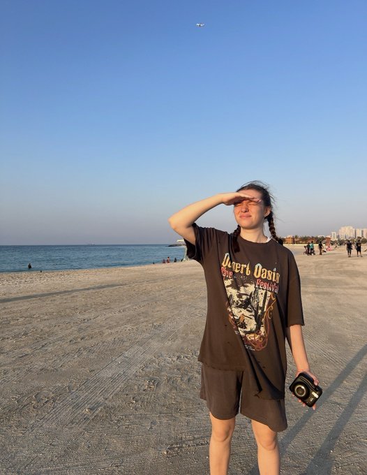 A photo of a young woman with braided hair standing on a sandy beach at sunset, wearing a 'Desert Oasis' t-shirt, holding a camera, and shielding her eyes from the sun.