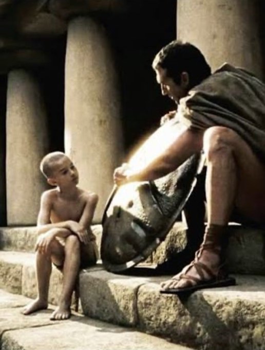 A young boy sits on stone steps looking up at a man holding a helmet, set against a backdrop of large classical columns reminiscent of ancient Roman architecture.