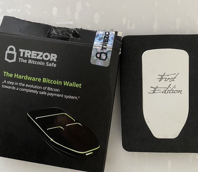A black Trezor hardware Bitcoin wallet box labeled "The Hardware Bitcoin Wallet" with its "First Edition" device shown next to it, featuring a holographic authenticity seal.