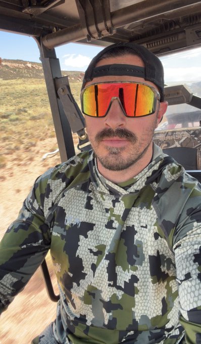 A selfie of a man wearing a backwards cap, reflective orange sunglasses, and a green and gray camouflage hoodie, seated in a utility vehicle with a desert landscape in the background.