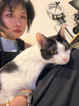 A young woman with short dark hair takes a selfie while holding a black and white cat with large yellow eyes, in an indoor setting with film equipment visible behind them.
